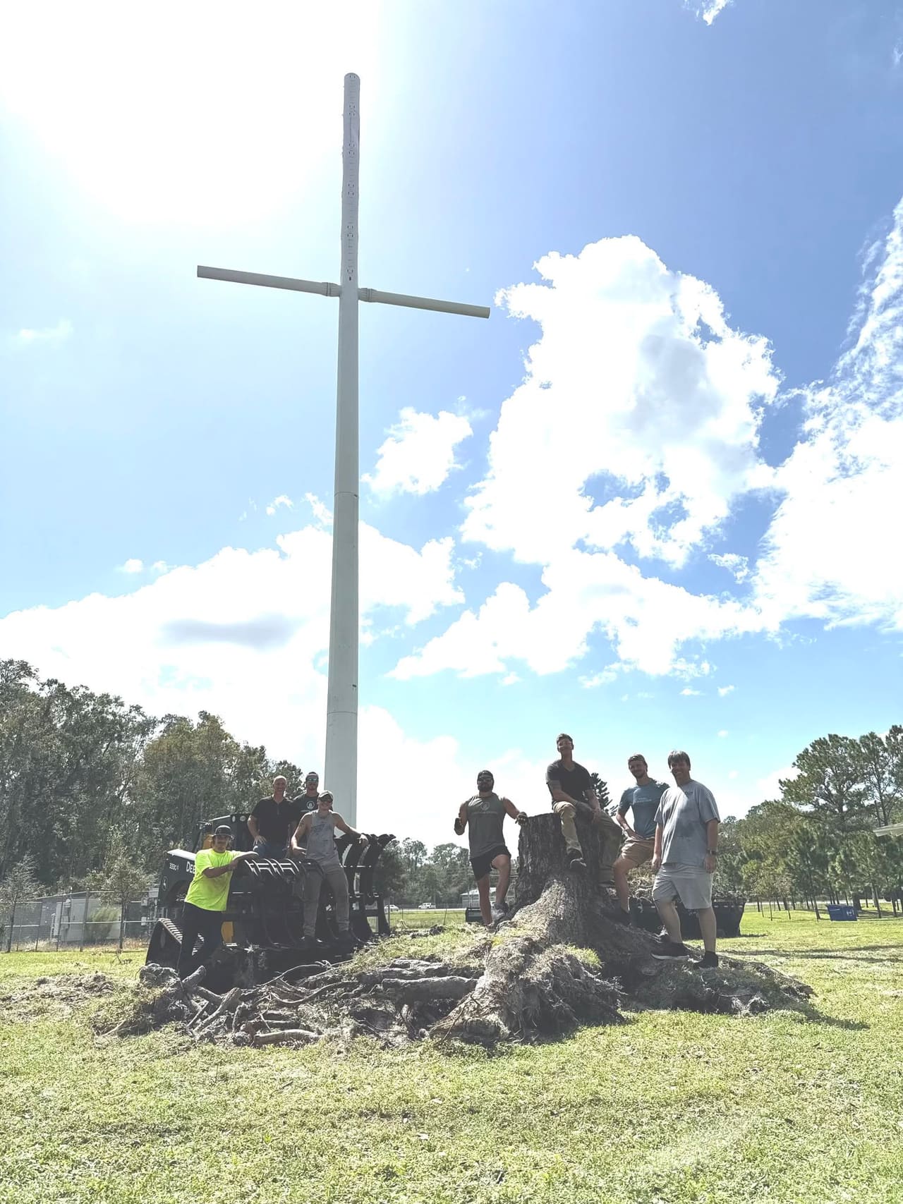 Eight people gathered around a large uprooted tree stump and compact equipment beneath a tall cross in a grassy outdoor area